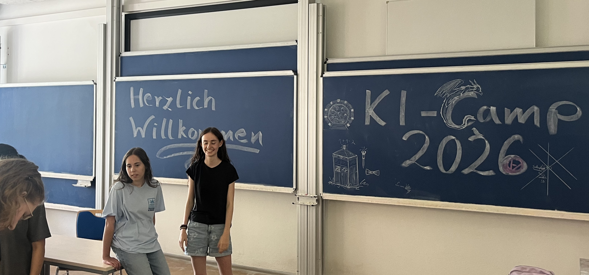 Schüler:innen KI-Camp 2026 Zwei Teilnehmerinnen vor einer Tafel mit der Aufschrift „Herzlich Willkommen – KI-Camp 2026“ in Hörsaal-125 des Instituts für Mathematik.