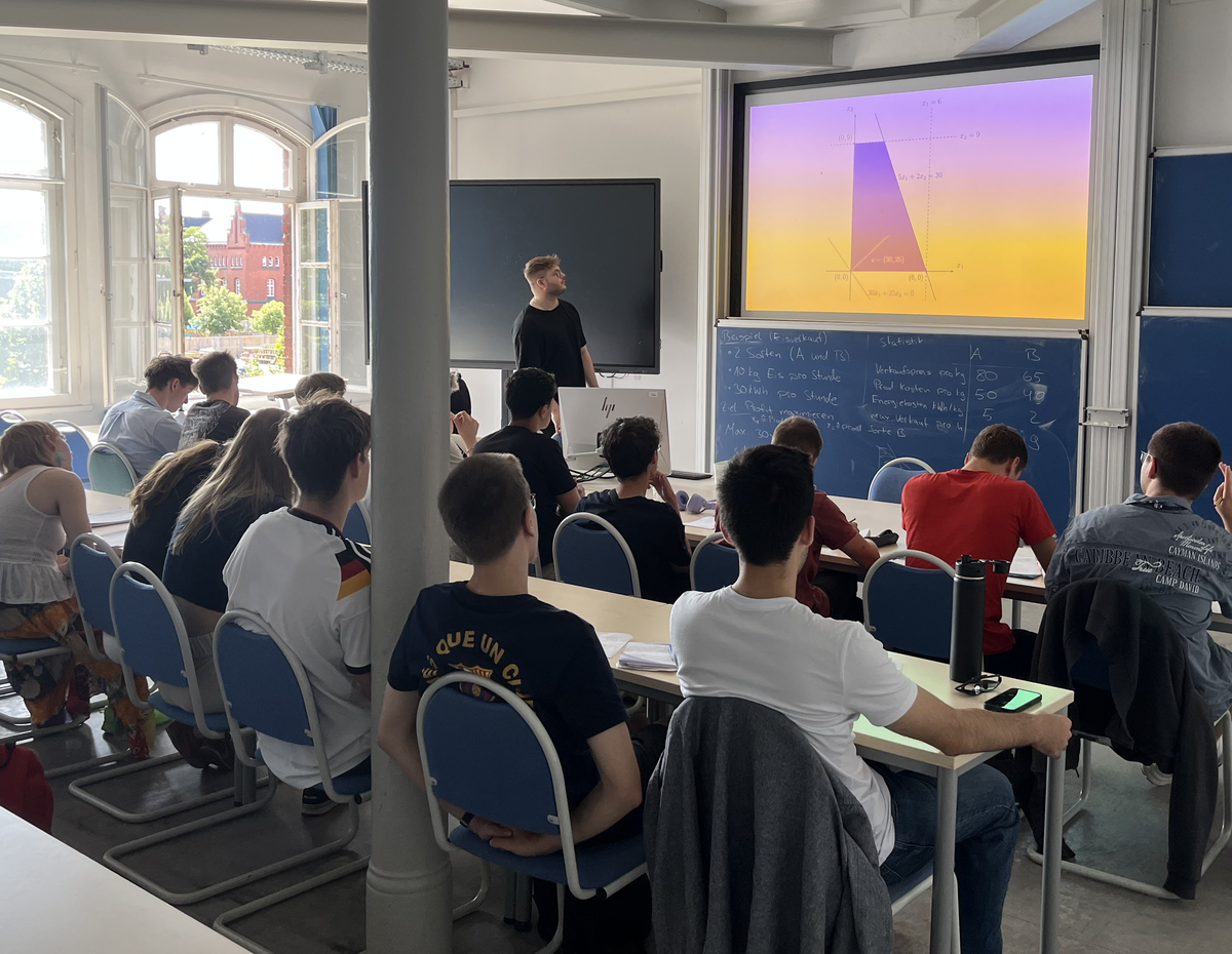Mathematik als Grundlage von KI Schüler:innen verfolgen in einem Seminarraum eine mathematische Erklärung von Cedric zu einem Optimierungsproblem auf einer Projektionsfläche.